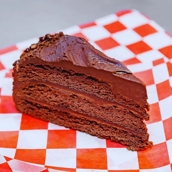 A rich slice of chocolate cake with multiple layers of frosting on red and white checkered paper.